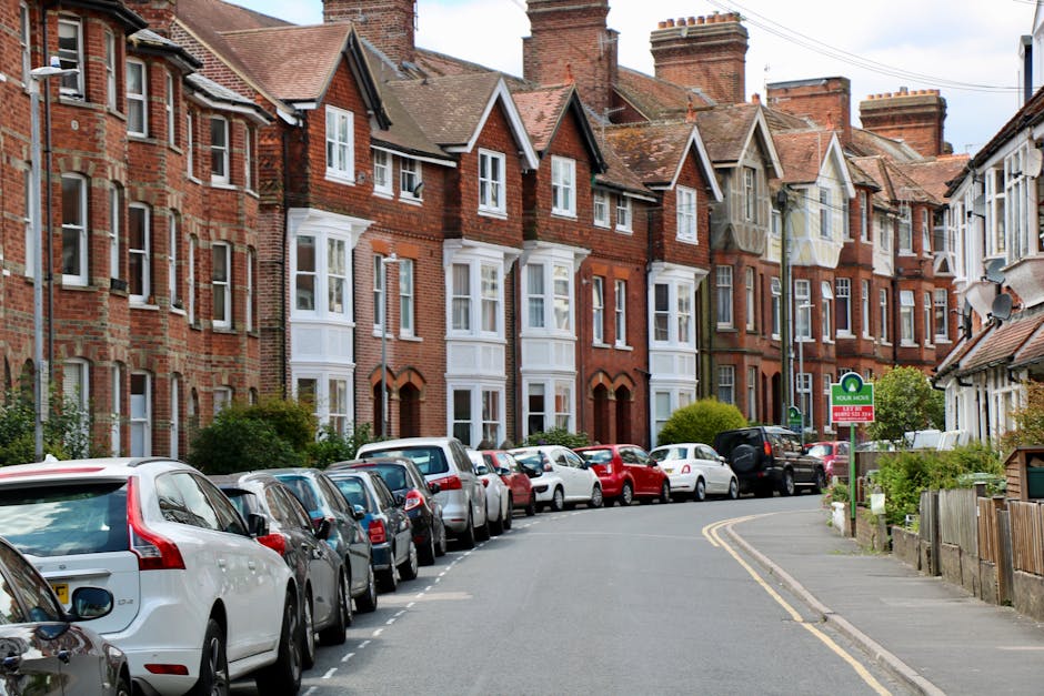 A residential street in Tufnell Park featuring a row of red-brick terraced houses with white window frames and small front gardens, some with greenery and shrubs. Several parked cars of various models